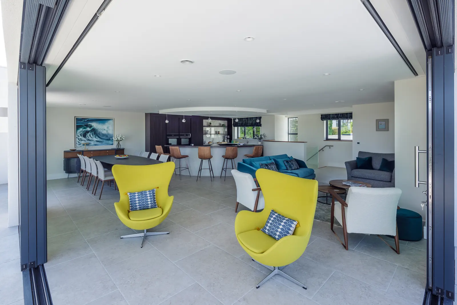 The first-floor living space with the bi-fold doors fully open onto the terrace, two yellow egg chairs in the foreground and the kitchen beyond